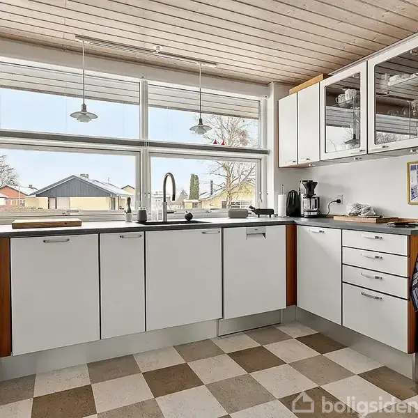 Køkken med hvide skabe og bordplade. Knapper og redskaber ovenpå. To loftlamper hænger over vasken. Gennem det store vindue ses en boligområde. Skakternet gulv og træloft.