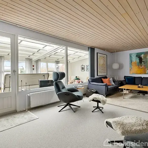 En stue med moderne møblering, herunder lænestole og sofaer arrangeret omkring et sofabord. Store vinduer og en glasskydedør giver udsigt til en overdækket terrasse. Væggene har kunstmalerier.