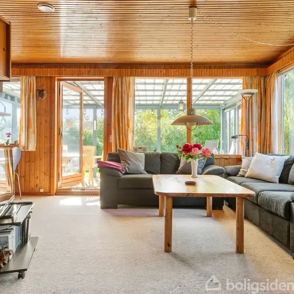 En hyggelig stue med en stor grå sofa, hvor der står en buket blomster på sofabordet. Rummet har trævægge og -loft og åbner ud mod en terrasse med grøn udsigt.