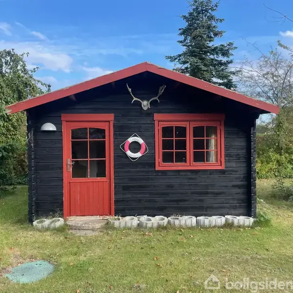 Et sort træhus med rød dør og vinduer står på en græsplæne, omgivet af buske og træer. En hjortekrans pryder husets facade, og en redningskrans hænger ved vinduet.