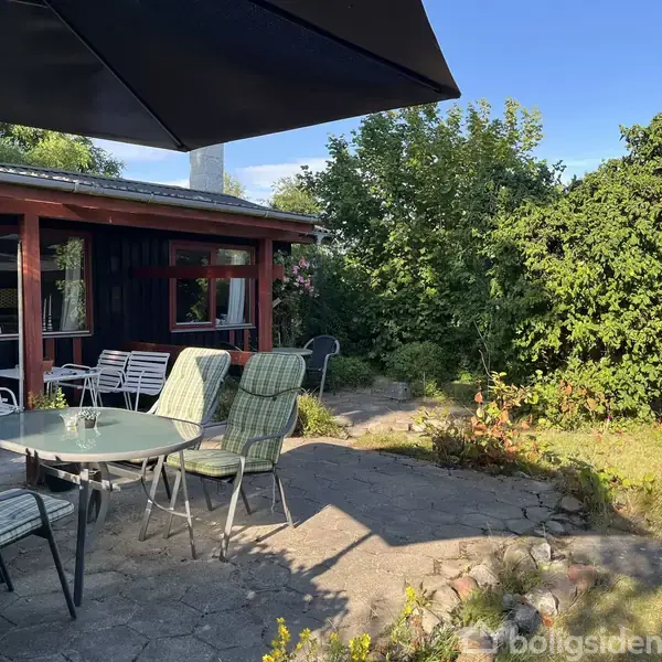 Terrassemøbler står på flisebelagt terrasseområde; en glasbord og polstrede stole under parasol. Ved siden af ses en sortmalet husfacade med store vinduer, omgivet af frodig have med buske og træer.