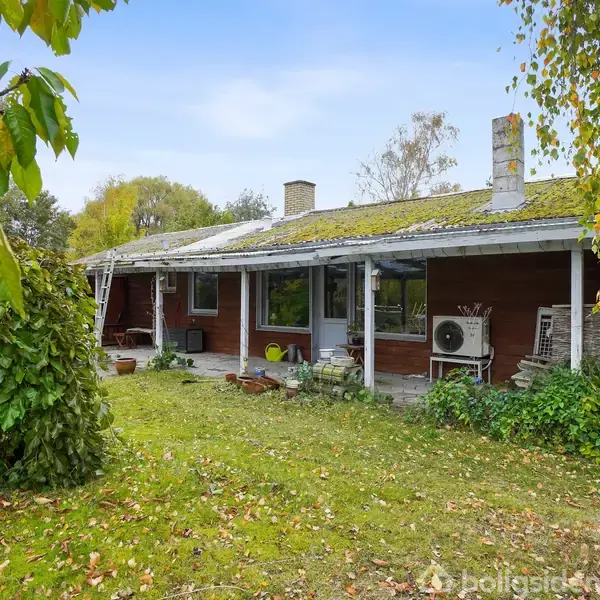 En rød træbygning med en enkelt vindue står i en have. Ved siden af er der en carport med et rødt stol. Baggrunden er fyldt med træer.