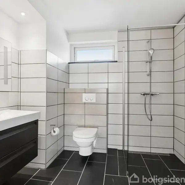 Badeværelse med væghængt toilet, bruser uden kabinet, hvidt rektangulært håndvaskmodul, og stort spejl. Væggene er i store hvide fliser, gulvet i sorte fliser. Rørformet håndklædetørrer til højre.