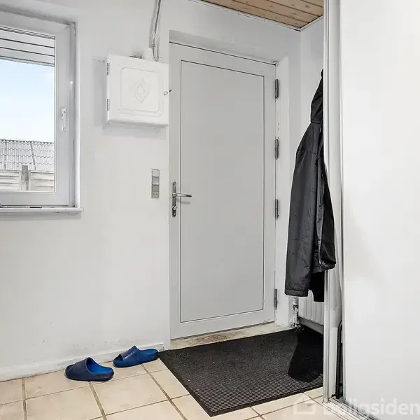 Dør står i en entré ved siden af vaskemaskine, med blå hjemmesko på flisegulv. Krog med jakker hænger ved siden af. En lys baggrund med vindue udenfor.