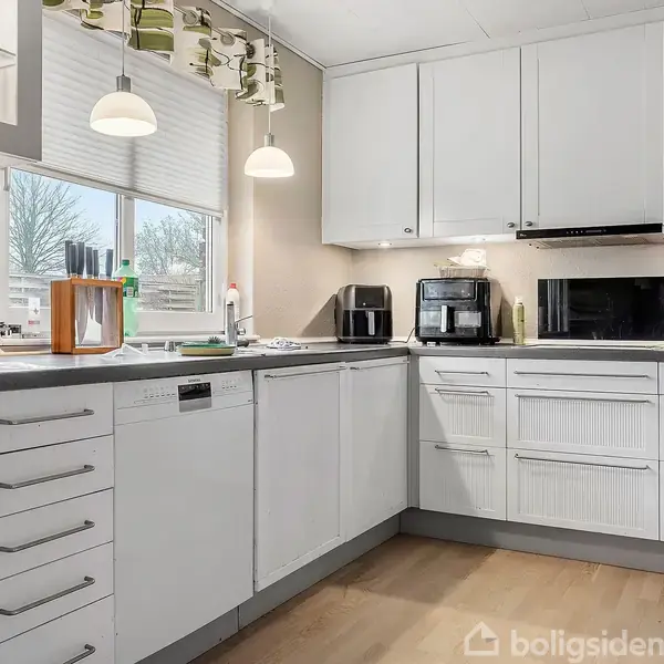 Et moderne køkken med hvide skabe og skuffer. Der er en opvaskemaskine, kaffemaskine og mikrobølgeovn. Lys træfarvet gulv med to hængende lamper over køkkenbordet. Vindue med udsigt.