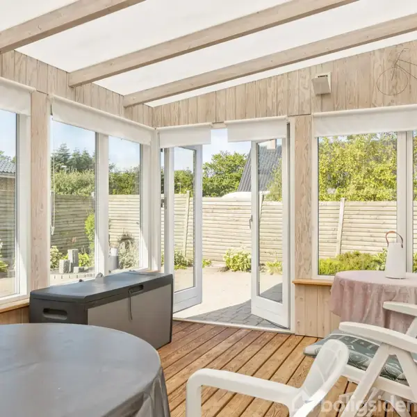 En lys havestue med træbeklædte vægge indeholder flere hvide stole omkring et bord. Dobbelt glasdøre åbner ud til en terrasse med træhegn og grønne planter.