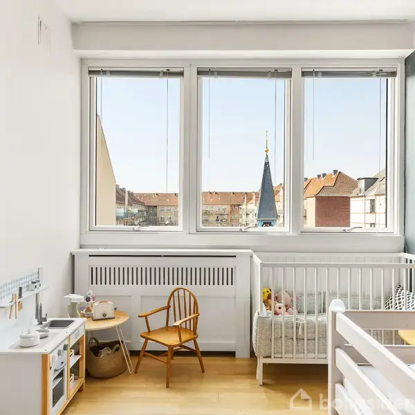 En lys børneværelse har en hvid tremmeseng, en lille træstol og et legetøjskøkken. Store vinduer giver udsigt til en by med tage og en kirketårn.