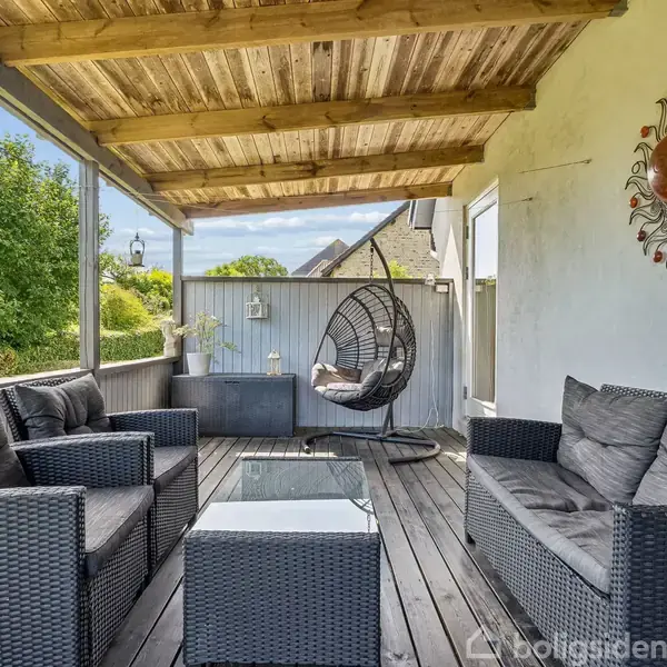 Terrasse med sorte kurvemøbler, sofaer omkring et firkantet glasbord. Hængende stol i hjørnet. Grønne omgivelser udenfor. Vægdekoration med en solformet metaludsmykning.
