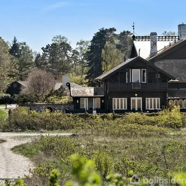 Et stort, sortmaleret hus med mørkt tag står i en frodig, skovklædt landskab. Huset har flere vinduer og balkoner, omgivet af græs og træer under en blå himmel.