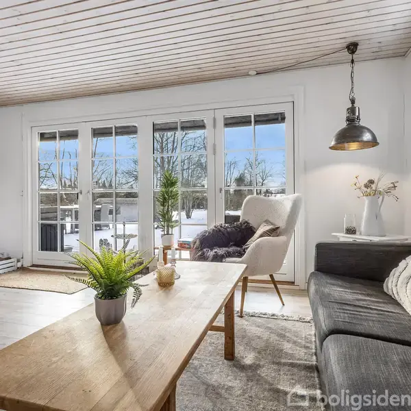 En stue indeholder en grå sofa, et træbord med en grøn plante og kopper, og en lænestol. Store vinduer med udsigt til en snedækket terrasse dominerer rummet.