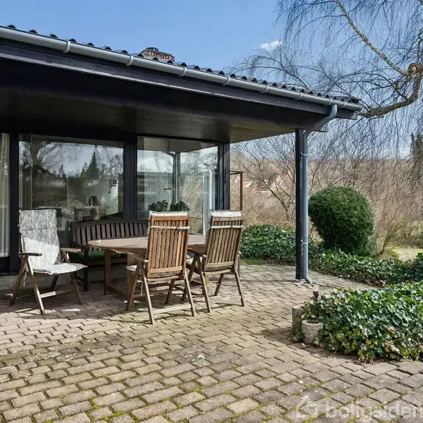En terrasse med havebord og stole står på en flisebelagt overflade. En mørk trætagkonstruktion giver skygge. Baggrunden viser græsplæne og træer under en klar blå himmel.
