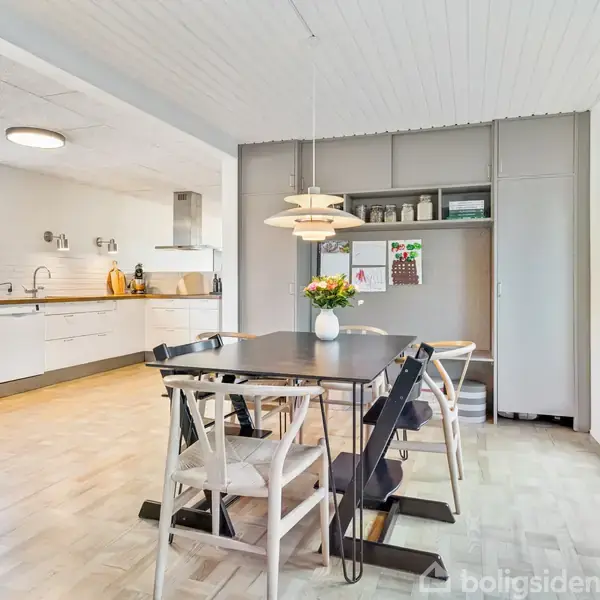 Et moderne køkken-alrum med et langt spisebord, omgivet af hvide stole, og en stor pendellampe. Baggrunden viser hvide køkkenskabe og apparater, med blomster på bordet.