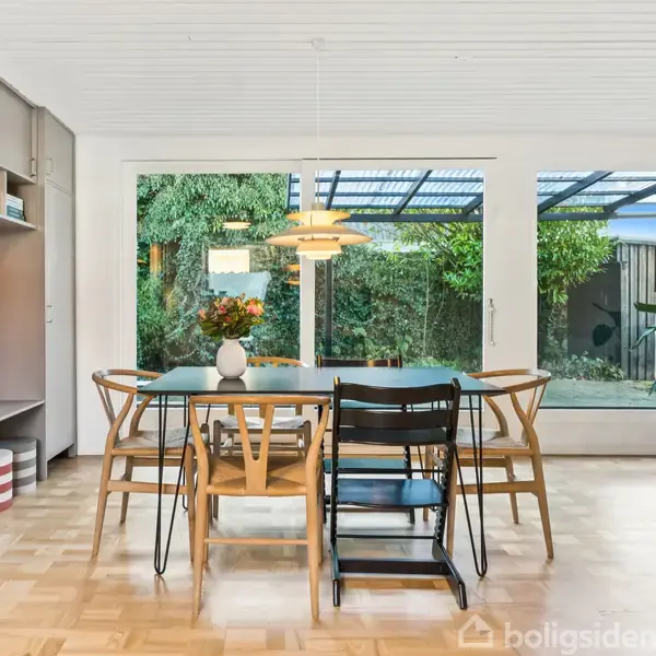 Et spisebord med stole står i et lyst rum med trægulv. Udenfor ses en grøn have gennem store vinduer, mens en loftslampe hænger over bordet.