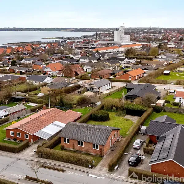 Byområde med røde murstenshuse omkranset af grønne hække set fra oven, nær kysten med udsigt over vandet. Husene er placeret langs veje med parkerede biler.