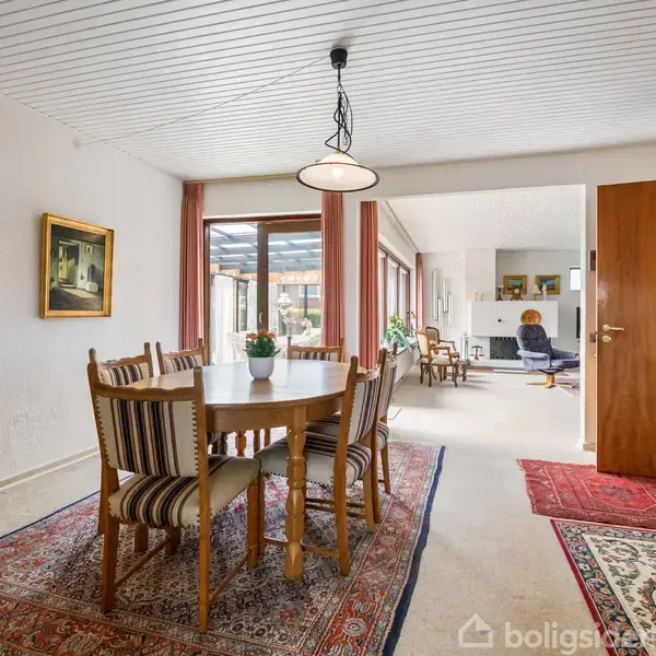 Et spisebord med seks stole står på et gulvtæppe i en stue. Rummet har hvide vægge, maleri, og gulvtæpper. Store vinduer bagved åbner op mod en lys stue.