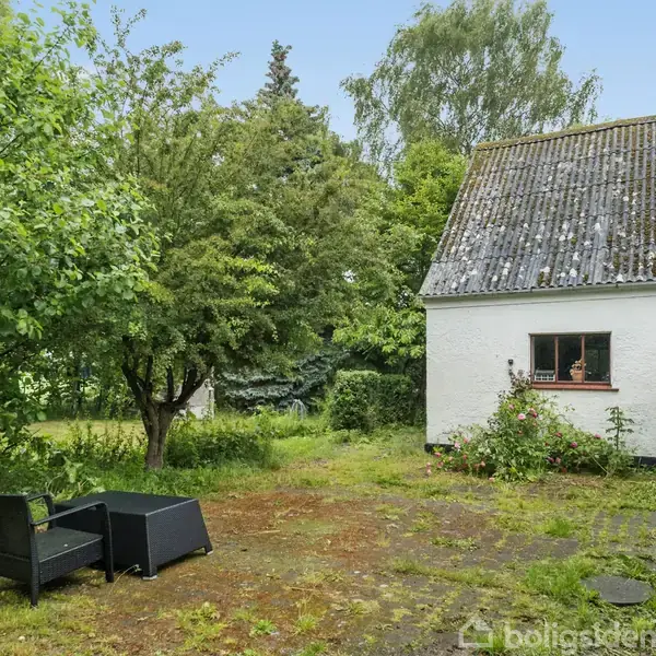 En ældre hvid bygning med mosgroet tag står på en gårdsplads. To sorte havemøbler er placeret på fliserne, omgivet af tæt grøn beplantning og træer.