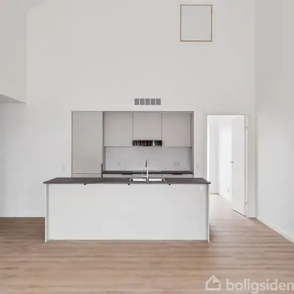 Et moderne køkken med en central ø og indbygget vask er placeret i et lyst rum med trælaminatgulv. Bag køkkenet er der en åben dør, der fører til et andet rum.