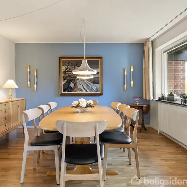 En spisebordsgruppe står placeret i en lyseblå væg-malet spisestue. Et rundt bord med otte stole er omgivet af kunst på væggene og vindue til højre. Bordet er pyntet med blomster.