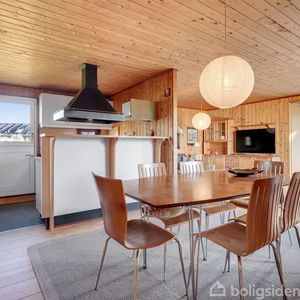 Et spisebord med seks stole står i et rum med trægulv og trævægge. Der er en lampe over bordet. Et åbent køkken ses i baggrunden.