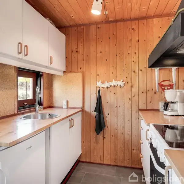 Et lille køkken med træpaneler på væggene, indeholdende en vask, hvide skabe, komfur, og kaffemaskine. Et viskestykke hænger fra en knagerække midt i rummet.