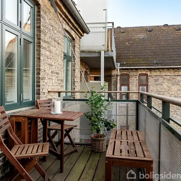 Et lille altanområde med et træbord og to stole, planter, og udsigt over et gammelt murstensbyggeri. Kaffen står på bordet, hvilket skaber en afslappet atmosfære.