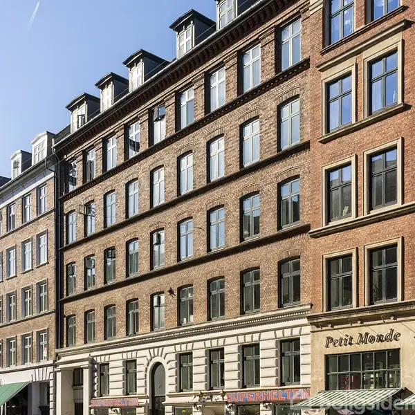 Et rødt murstensbygning har flere vinduer på hver etage med butikker i stueetagen. Tekst på butikkerne omfatter "Petit Monde" og "Legetøj". Klar himmel i baggrunden.