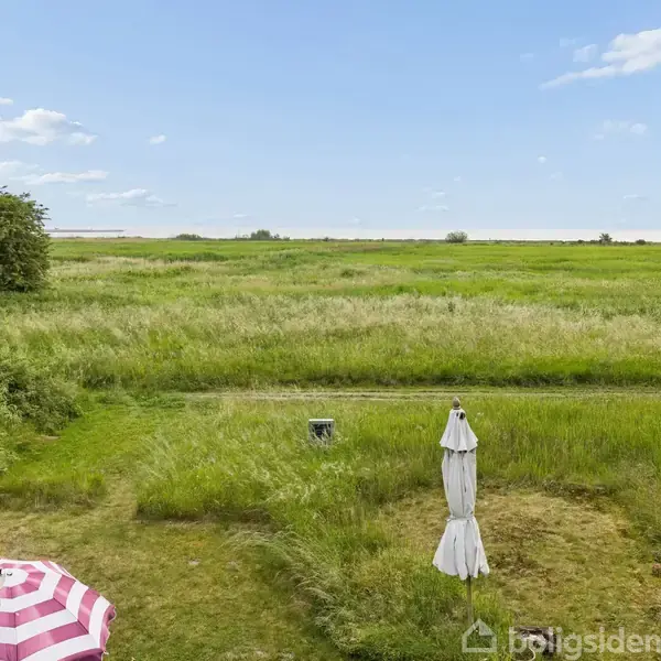 En parasol står foldet sammen i en åben græsmark. I forgrunden er der en terrasse med en lyserød-hvid stribet parasol. Baggrunden viser vide grønne marker under en blå himmel.