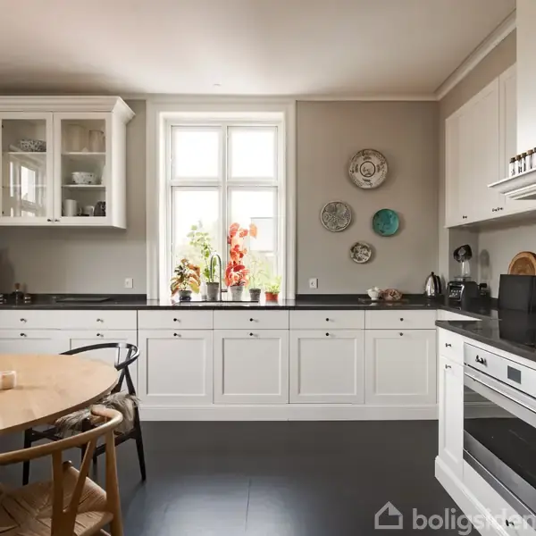 Et hvidt køkken har skabe, komfur og et rundt spisebord med stole. Vinduet midt i rummet lader naturligt lys ind, mens dekorationer pryder væggene.