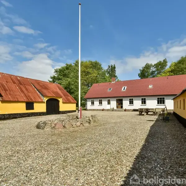 Gårdbygninger omkring en grusbelagt gårdsplads med en flagstang centralt. Gule vægge og røde tage omkranser gårdspladsen. Træer i baggrunden skaber en landlig atmosfære.
