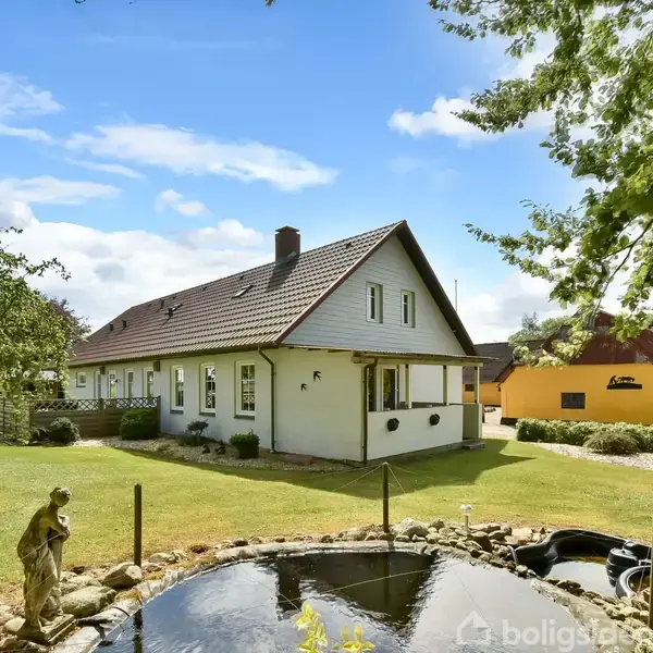 Hvidt hus med skråt tag står stille ved siden af en stor have. Foran huset er en lille dam omkranset af sten. Træer og græs omgiver området på en solrig dag.