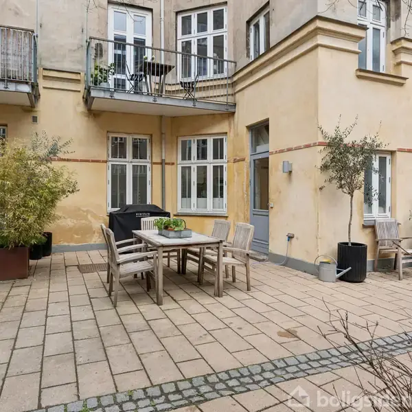En gårdhave med et bord og stole står på en flisebelagt terrasse, omgivet af planter i krukker, cykler og en historisk bygning med altaner i baggrunden.