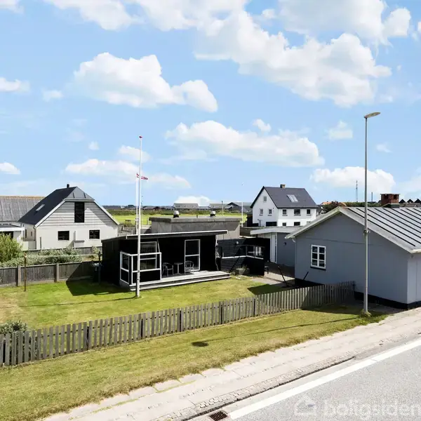 Et sort sommerhus med flagstang står på en velplejet græsplæne omgivet af et træhegn. Flere huse ses i baggrunden under en blå skyet himmel.