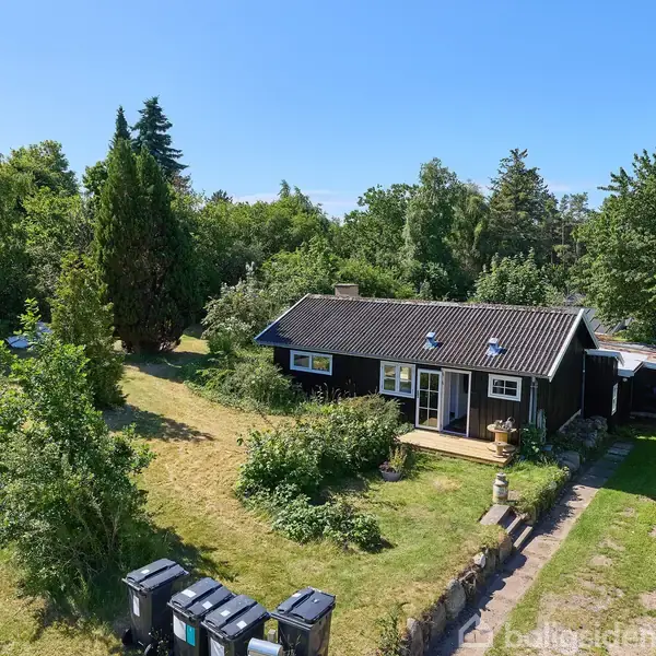 Et sort sommerhus med hvide vinduer står i en frodig have omgivet af træer og buske. Foran huset er der en græsplæne og affaldsbeholdere.