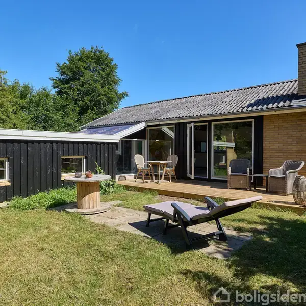Et moderne hus med terrasse, hvorpå der står stole og bord, ligger i en frodig have. En træbænk står på græsplænen. Baggrunden er lys og grøn af buske og træer.
