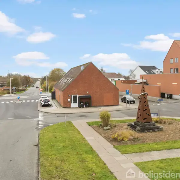 En skulptur står på en græsplæne midt i en rundkørsel. Baggrunden indeholder røde murstensbygninger, veje med biler og en blå himmel med skyer.