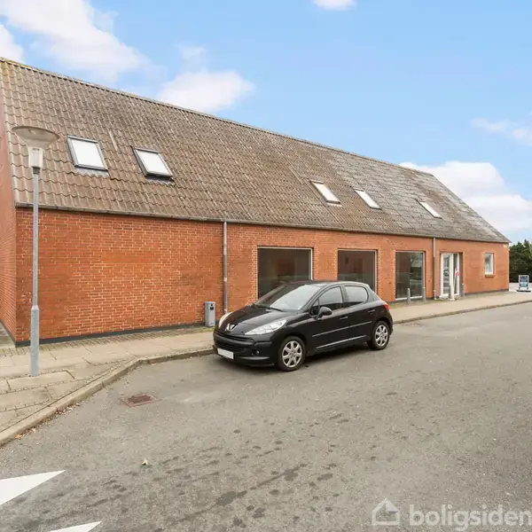 En sort bil holder parkeret foran en lang, rød murstensbygning med skråt tag og tagvinduer. Bygningen ligger på et gadehjørne i en byzone med fortove og vejskilte.
