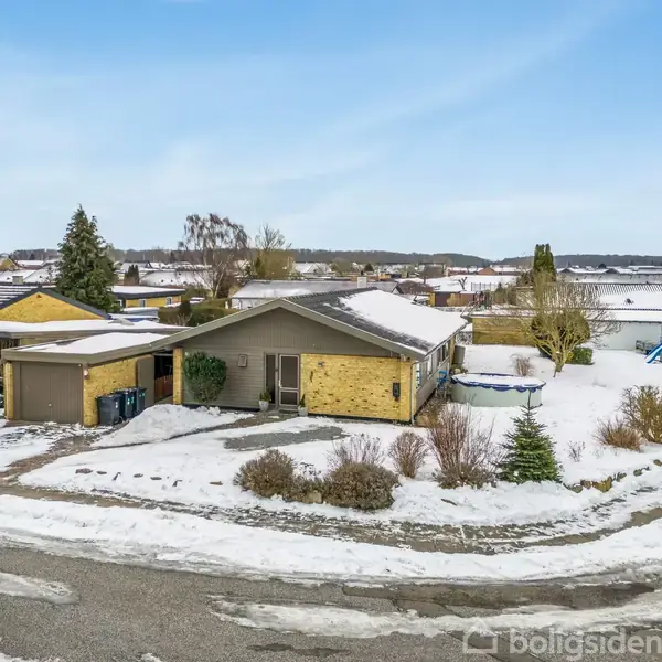 En enfamilieshus er beliggende i et snedækket boligområde. Huset har en gul murstensfacade og tilstødende garage. Omgivelserne inkluderer sneklædte tage og bare træer.