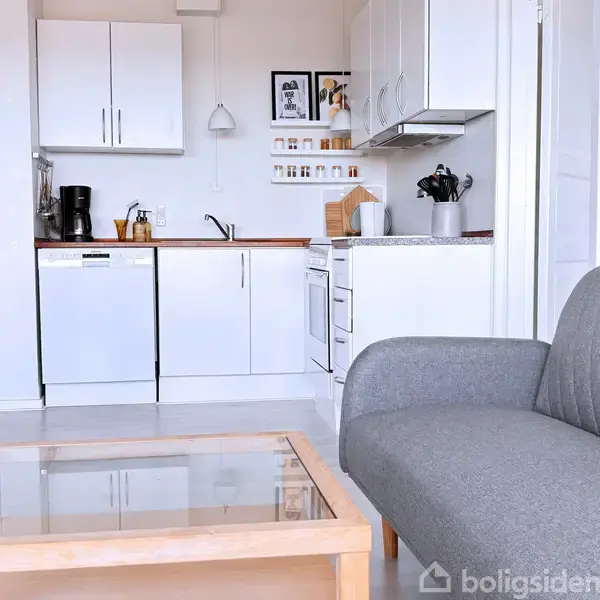 En grå sofa står foran et glas-sofabord i en stue med hvidt tekøkken. Køkkenet har en opvaskemaskine, kaffemaskine, og hylder med krydderier. Garderobe og entre ses i baggrunden.