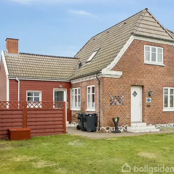 Rødt murstenshus med gråt tag, flankeret af mindre bygning og træhegn. Stående på en grøn plæne med blå himmel i baggrunden. Ingen synlig tekst i billedet.