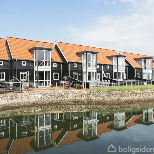 Rækkehuse med sorte facader og orange tage står langs en kanal. Havnefrontmiljø giver reflekser i vandet, og boligerne har store glaspartier, der giver udsigt til det omkringliggende område.