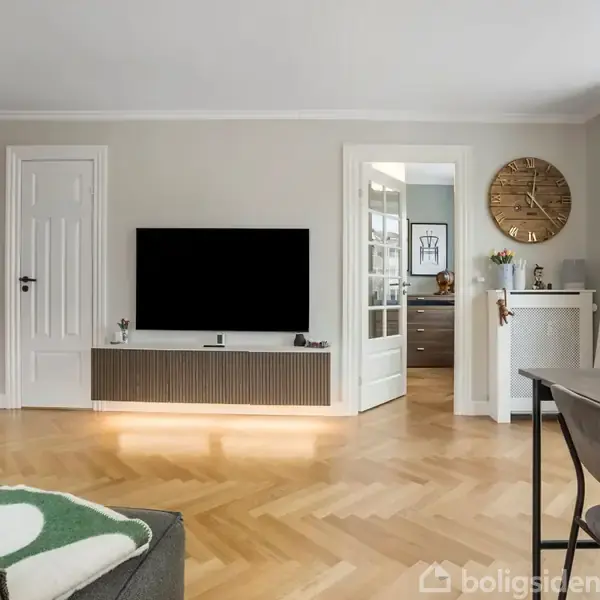 TV hænger på en væg over en moderne skænk i en stue. Til højre er der en stor ur, plante og lys indstrømning fra vindue.