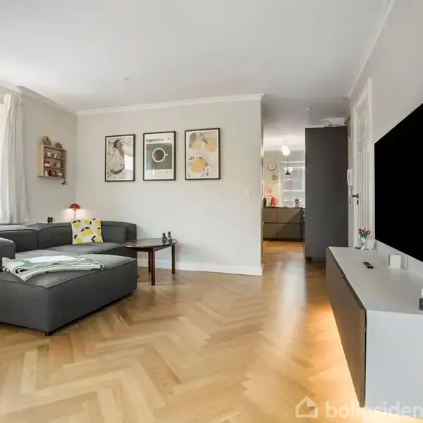 Gråt sofaarrangement står stille i et lyst stueområde med parketgulv; vægudsmykninger og malerier indrammer rummet, mens et fjernsyn er placeret på en hvid skænk i baggrunden.