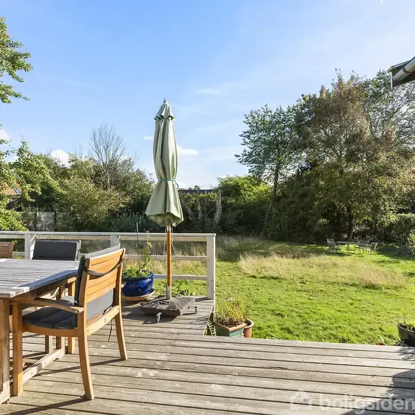En træterrasse med et bord og stole står foran en stor, græsbevokset have omgivet af træer. En parasol er lukket og placeret ved siden af bordet.