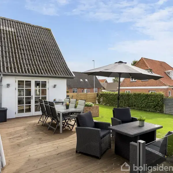 Træterrasse med spisebord, stole og parasol står foran et hvidt hus med glasdøre. Omkring terrassen er der en grøn græsplæne og nabohuse med røde tage.