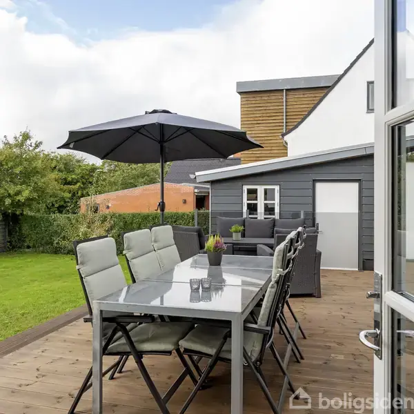 En moderne terrasse har et spisebord med flere stole og en stor parasol. Bordet er dekoreret med vaser, og terrassen er omgivet af en velplejet grøn have og et hus i baggrunden.