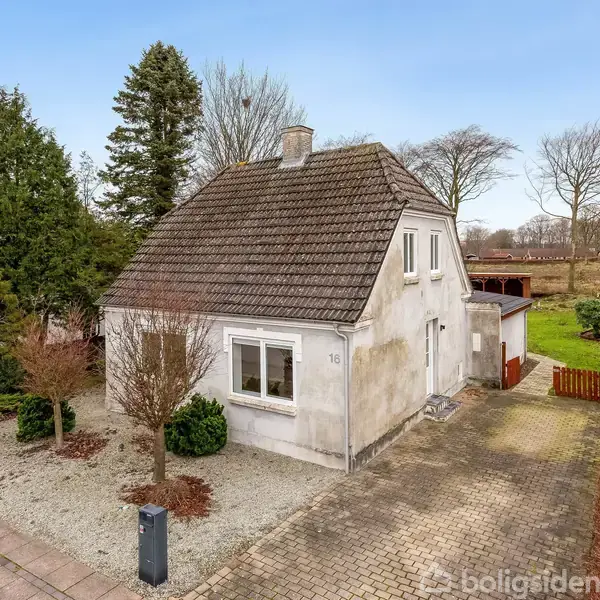 Et hvidt hus med gråt tegltag står på en grusbelagt grund. Det er omgivet af træer og har en brostensbelagt indkørsel. Husnummeret 15 er synligt på facaden.
