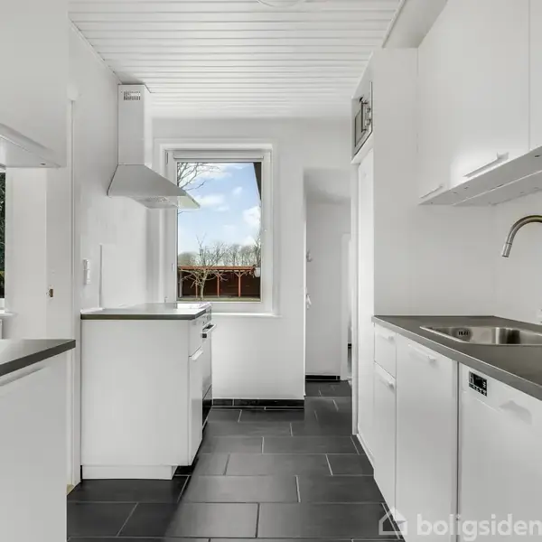 Moderne køkken med hvide skabe, sort bordplade og stålvask. Rummet har mørke fliser og et vindue med udsigt til træer, hvilket giver lysindfald. Vægge og loft er hvide.