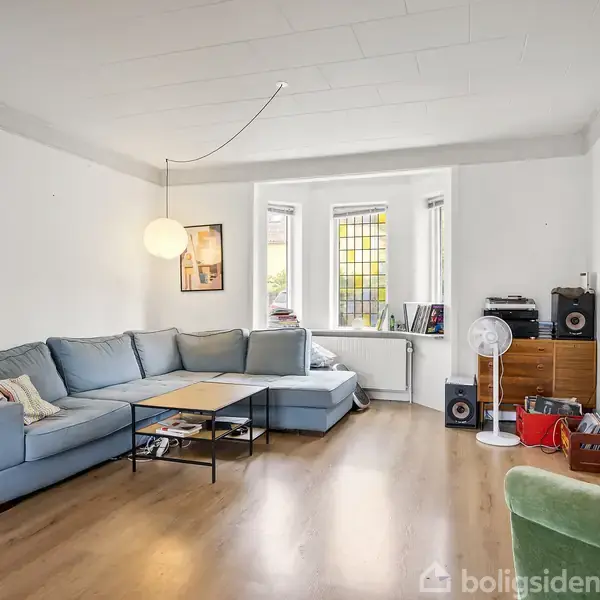 Blå sofagruppe står mod en hvid væg i en stue med trægulv. Et sofabord er foran sofaen. Rummet har store vinduer, planter, og en stående lampe. Flere bøger ligger på gulvet.