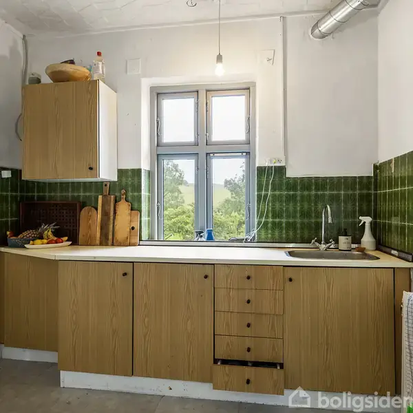 Køkkenskabe med trælook monteret på væggen, et vindue i midten ovenover vasken med udsigt til træer. Grønne fliser dækker halvt væggene. Rengøringsartikler og skærebrætter står på bordet.