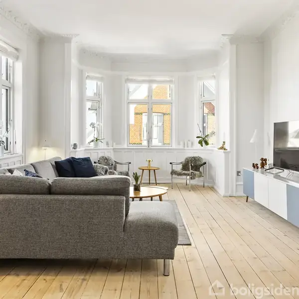 Grå sofa står ved et sofabord i en lys, rummelig stue med trægulve. Vinduerne slipper lys ind, og en tv-bænk står overfor sofaen med en stor skærm.